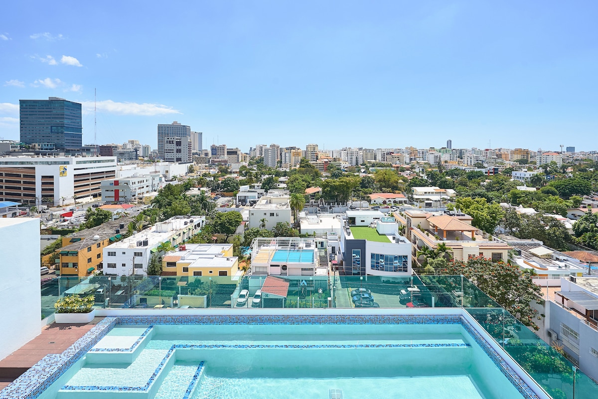 A rooftop terrace showcases a large pool with clear blue water, offering expansive views of the cityscape. Surrounding greenery and contemporary buildings are visible, under a clear blue sky. The terrace layout is framed by glass railings, enhancing the scenic atmosphere.