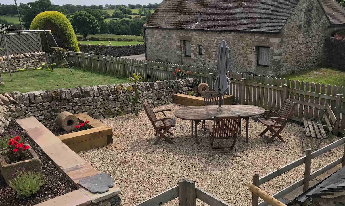 An outdoor patio area is displayed, featuring a circular wooden table surrounded by several wooden chairs. Gravel covers the ground, complemented by colorful flower beds. A shaded umbrella stands at the center of the table, while a stone wall and wooden fence frame the space. The backdrop includes rolling hills.