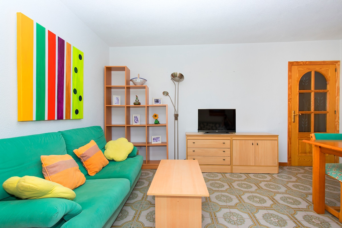 A living area is depicted with a green sofa adorned with colorful cushions, complemented by a light wooden coffee table. A shelving unit displays decorative items, while a television is positioned on a matching wooden cabinet. The floor features patterned tiles, adding visual interest.