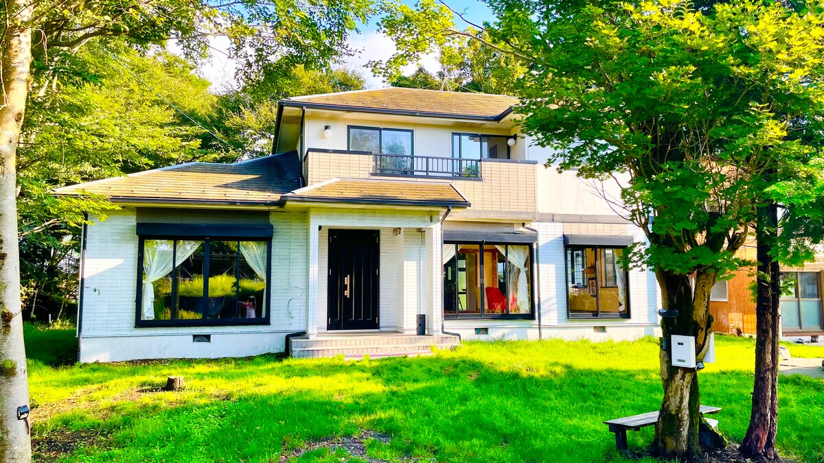 The two-story house is situated in a lush green setting, featuring large windows that allow natural light to illuminate the interior. A welcoming entrance leads to the front porch, while a mix of trees and grass surround the property, enhancing its serene atmosphere.