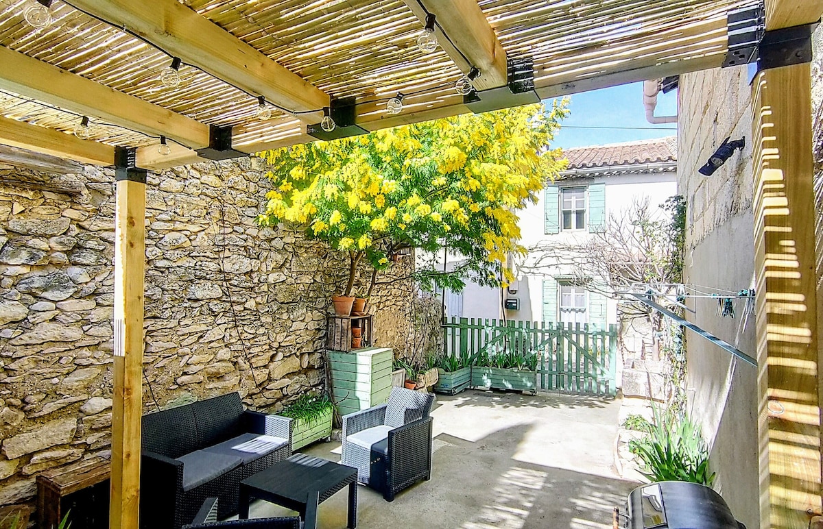 An outdoor area is shaded by a wooden pergola, featuring a seating arrangement with dark furniture. A vibrant yellow tree provides natural greenery, while stone walls add rustic charm. A small fence and potted plants create an inviting atmosphere for relaxation or dining.