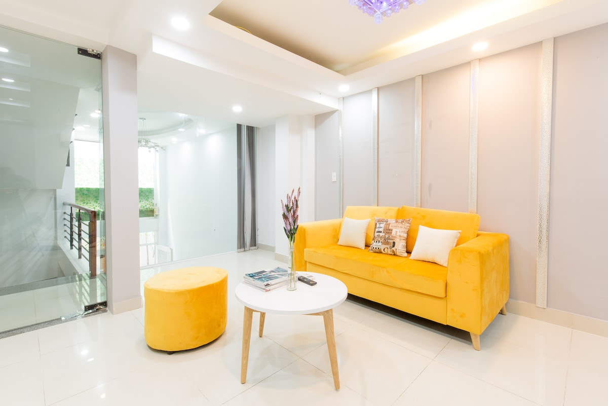 A modern living area features a bright yellow sofa paired with a round ottoman. A small coffee table is centered in front, adorned with a magazine and a vase of flowers. The walls are light-colored, creating a fresh and inviting atmosphere.