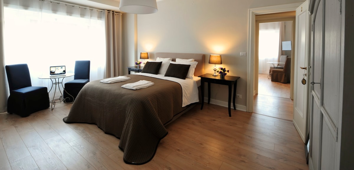 A bright bedroom features a spacious bed with neutral-colored bedding. Two bedside lamps illuminate the room, while a small dining table and chairs are positioned nearby. Natural light streams in through large windows, and a doorway leads to an adjoining room.