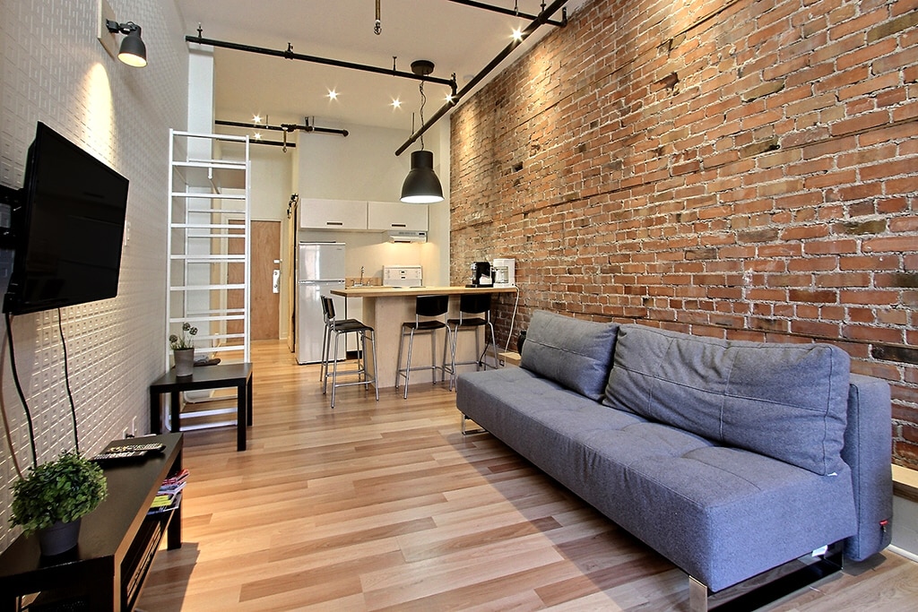 The spacious living area features exposed brick walls and wooden flooring. A modern gray sofa is positioned across from a wall-mounted television. A dining area with high stools and a kitchen visible in the background provides a contemporary, open layout.