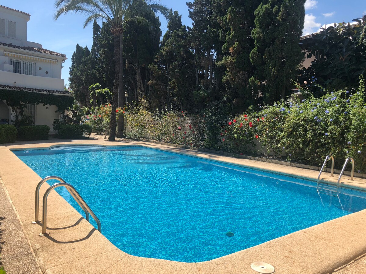 A bright swimming pool is surrounded by lush greenery and flowering plants, providing a refreshing space. A set of metal steps leads into the clear blue water, while palm trees offer shade along the edges, creating a serene outdoor setting within the community.