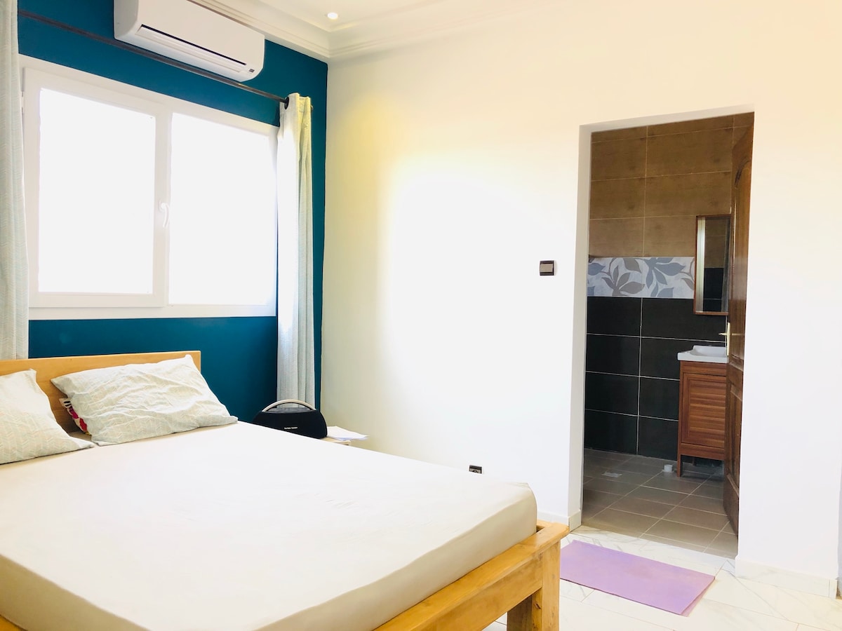 A bright and quiet bedroom features a spacious wooden bed with light bedding. Natural light enters through two large windows, and a bathroom entrance is visible in the background. The walls are painted in a calming blue tone, contributing to a serene atmosphere.