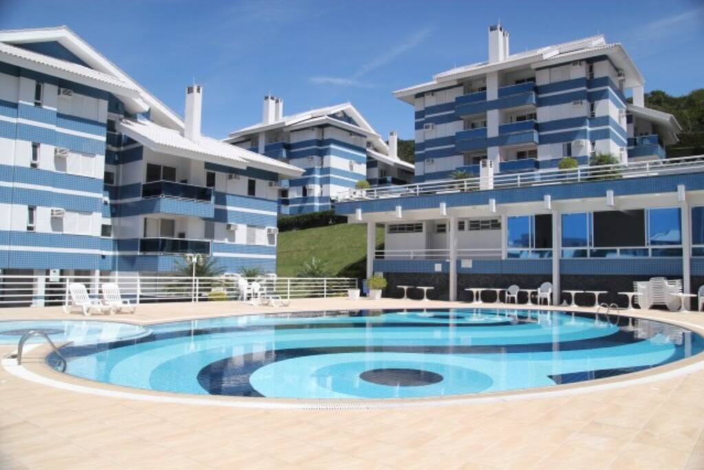 Piscina do condomínio com salão de festas e vestiários ao fundo.