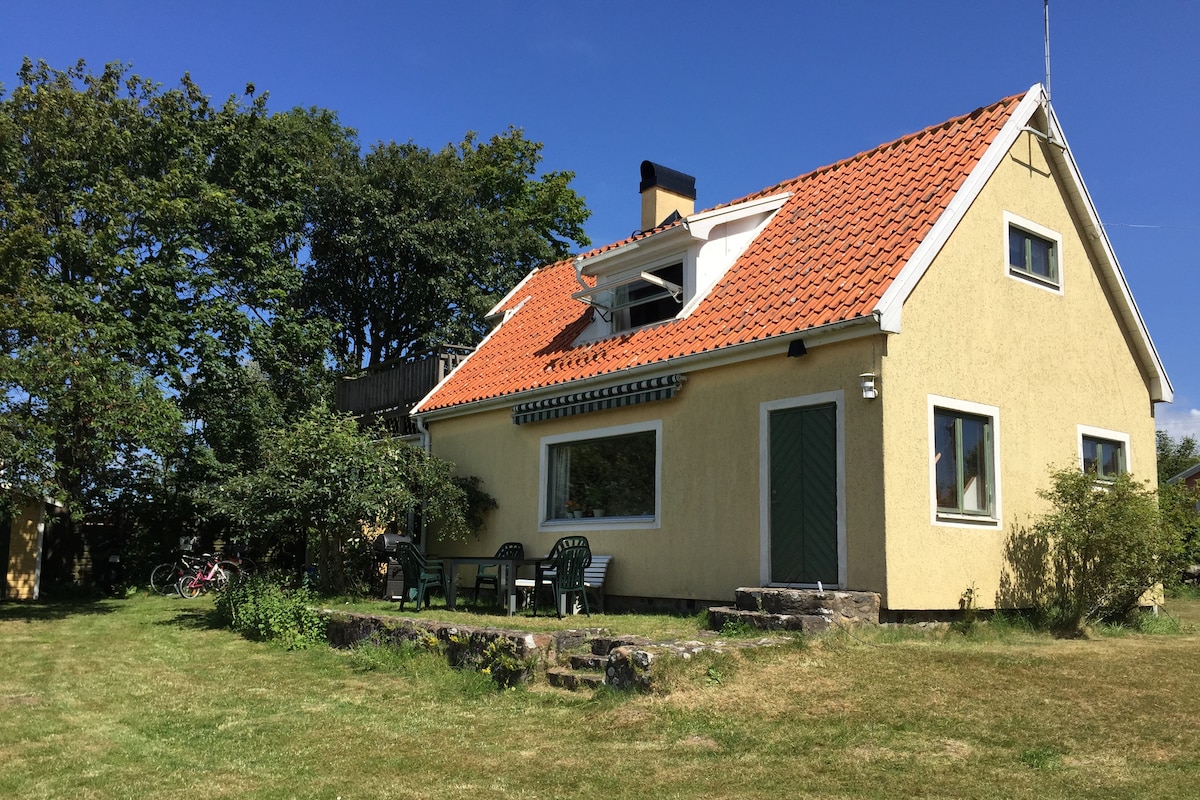 The house features a yellow exterior with a sloped red-tiled roof, surrounded by greenery. A stone staircase leads to a patio furnished with several chairs. A bicycle can be seen beside the house, and a spacious lawn invites outdoor activities.