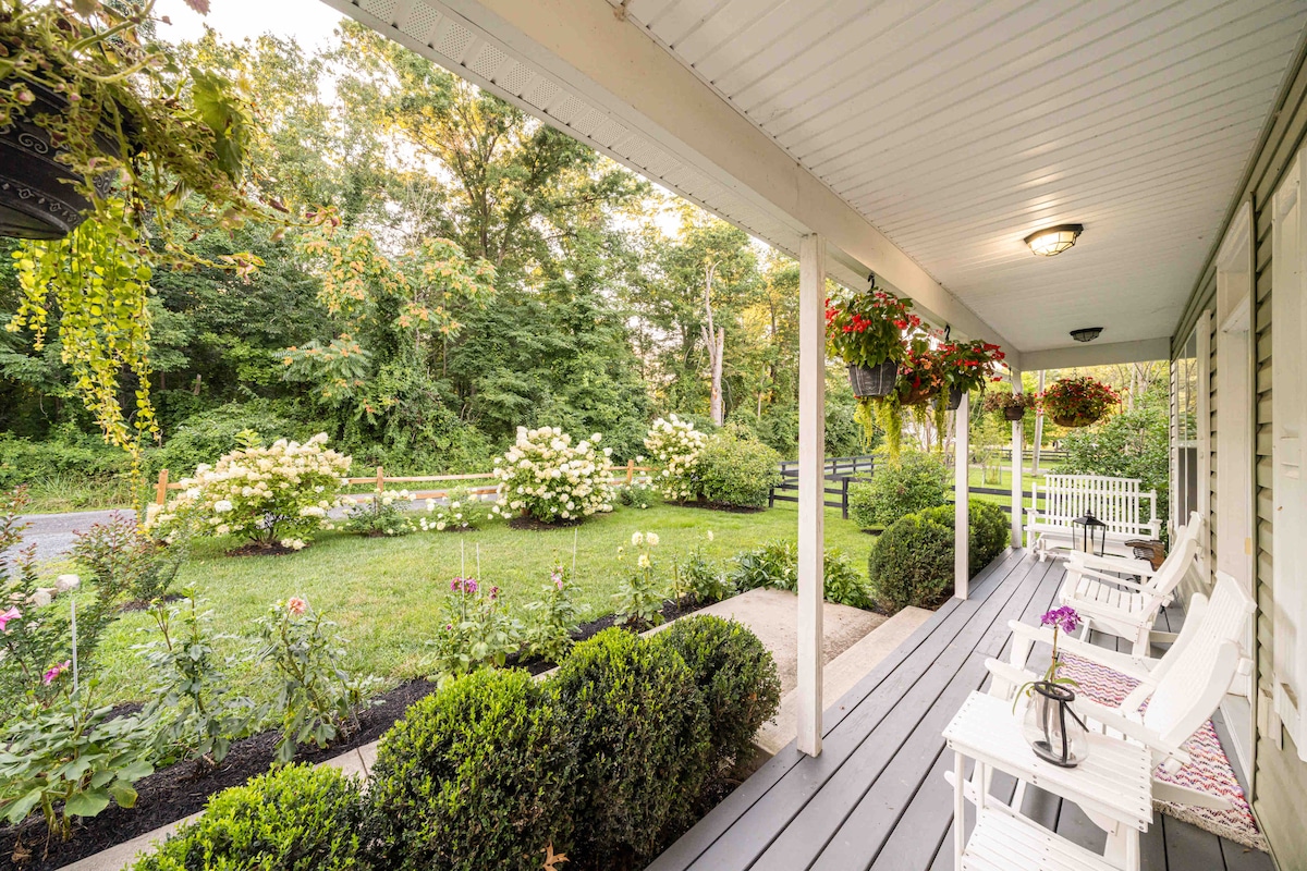 The front porch features white Adirondack chairs and a small table, inviting relaxation. Hanging planters add greenery, while nearby gardens showcase blooming flowers. A peaceful view of the surrounding trees and well-maintained yard is visible, providing a serene backdrop.
