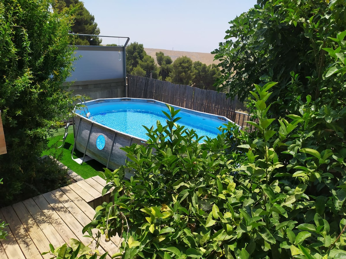 A round pool is set within a wooden deck, surrounded by lush greenery. The clear blue water reflects the sunlight, while a wooden fence offers a sense of privacy. Bright green grass features beneath the pool, enhancing the calming natural environment.