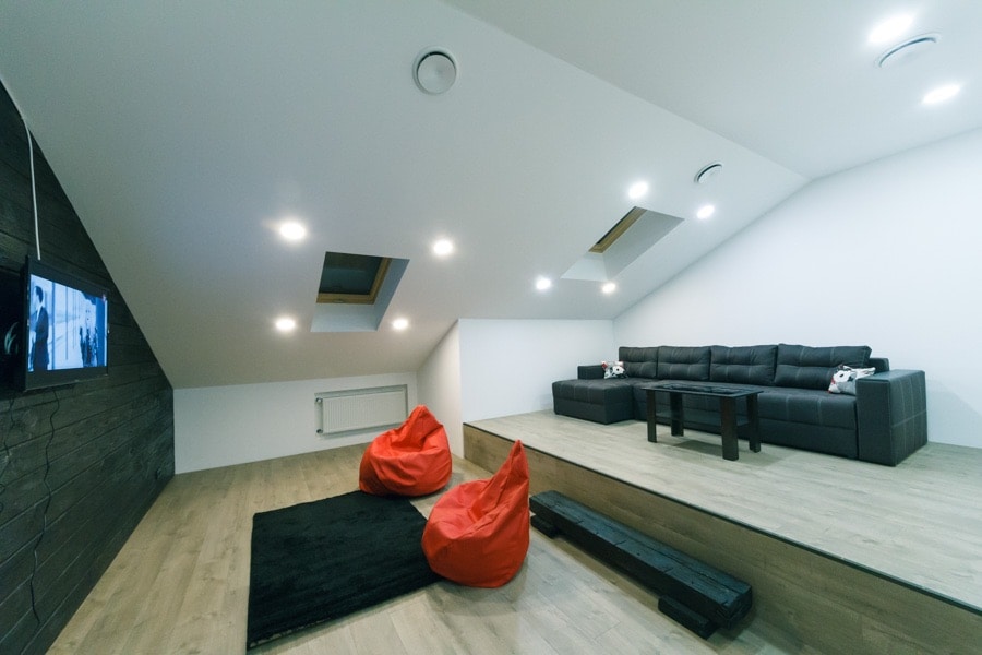 An attic space features a stylish dark gray sofa facing a mounted flat-screen television. Two red bean bag chairs are positioned on a soft black rug. A raised platform adds dimension, while skylights allow natural light to enter, enhancing the contemporary design.