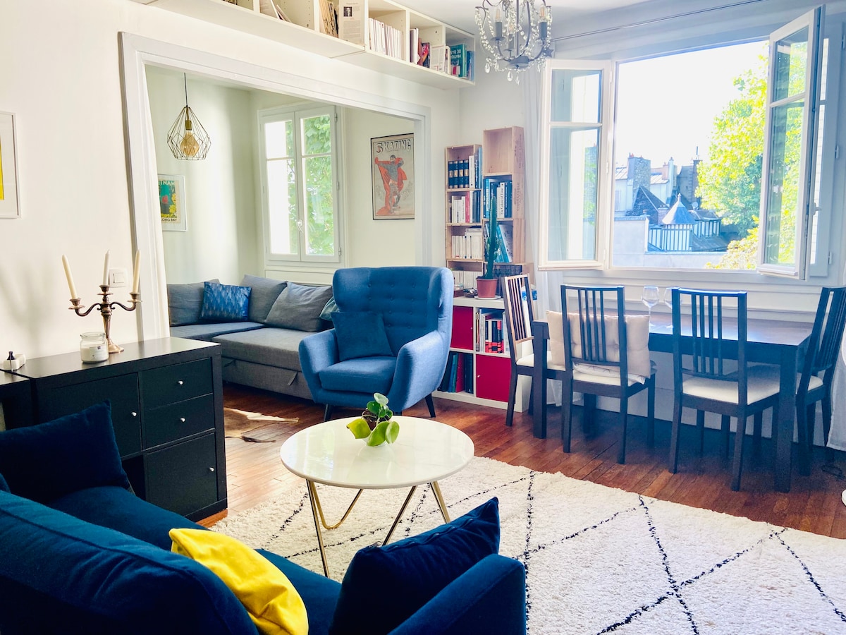 A spacious living area is illuminated by natural light from large windows. A comfortable blue armchair and a cozy sofa are arranged around a round coffee table. A dining set for four is visible, alongside bookshelves filled with various books.