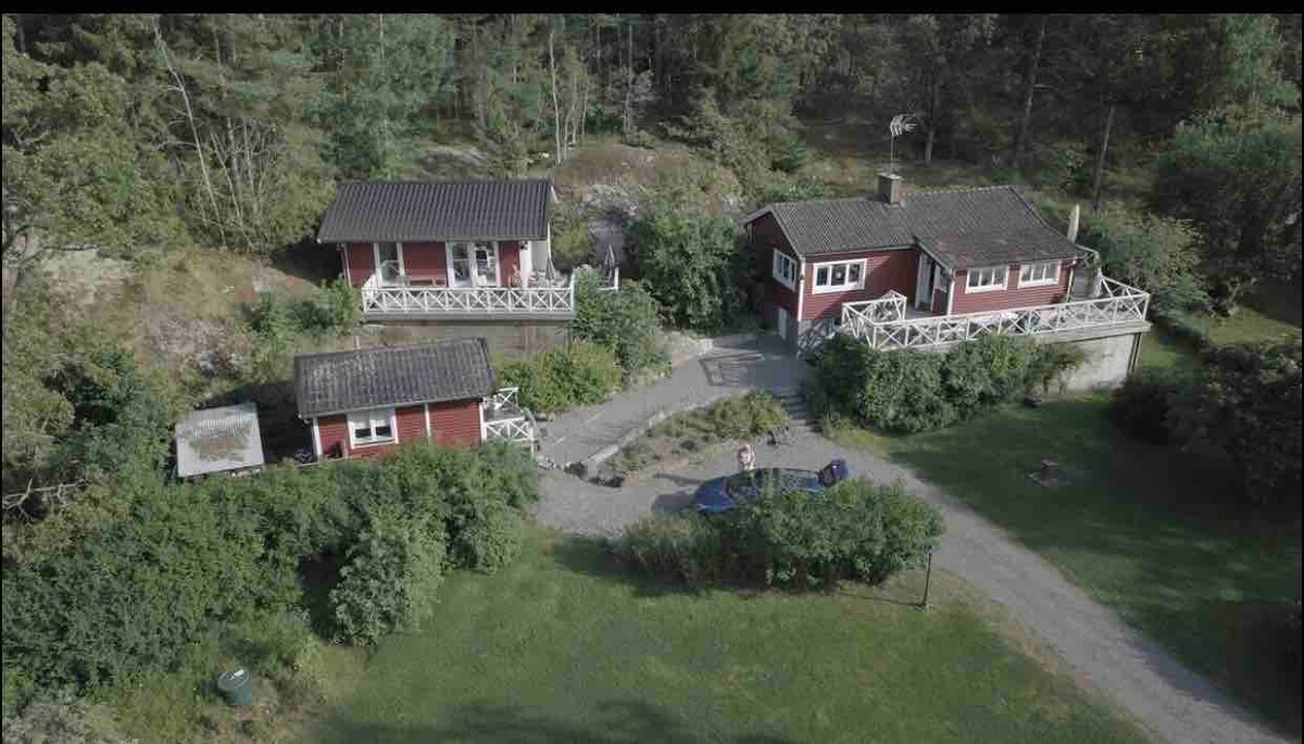 Three charming red houses are arranged in a natural setting, surrounded by greenery. A gravel pathway connects the homes, which feature covered porches. The lush lawn offers ample outdoor space amidst trees, creating a serene atmosphere. The image captures the proximity of the houses to each other.