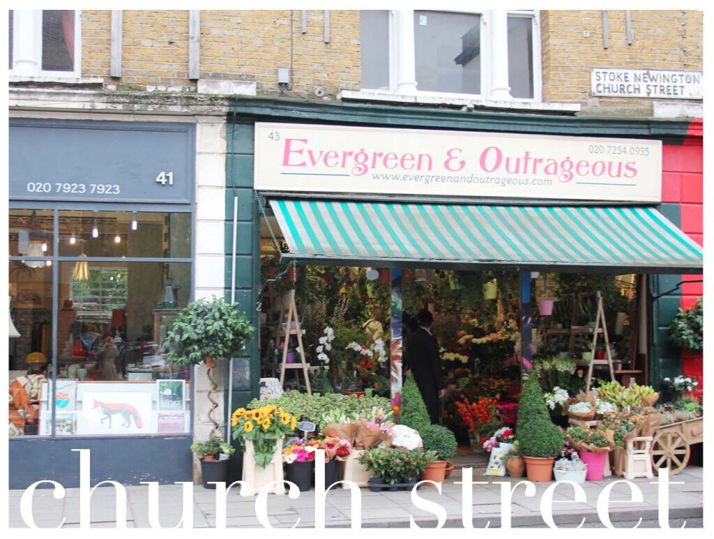 A vibrant storefront named 'Evergreen & Outrageous' is displayed, featuring a variety of potted plants and flowers arranged at the entrance. Multicolored flower pots line the sidewalk, and a striped awning provides shade over the shop. A person can be seen tending to the plants.