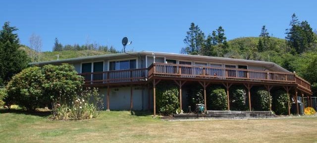 A single-story home with a broad wooden deck extends along the front, providing ample outdoor space. The building is surrounded by well-maintained greenery and features large windows that invite natural light. A grassy area is visible in the foreground, creating a serene outdoor setting.