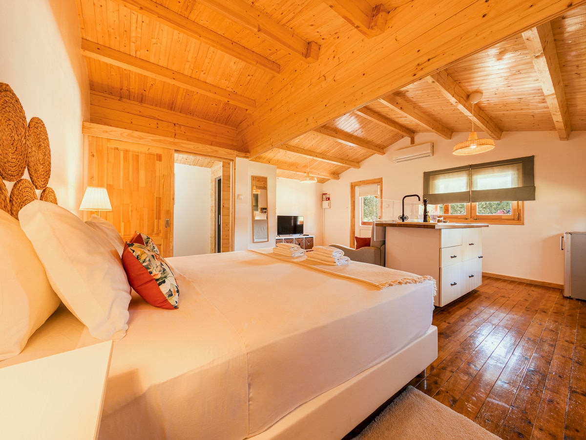 The bright interior features a spacious sleeping area with a large bed adorned with soft linens and decorative pillows. Natural wood beams create a warm ceiling above. An open living space includes a kitchenette with modern amenities, a seating area, and windows allowing ample natural light.
