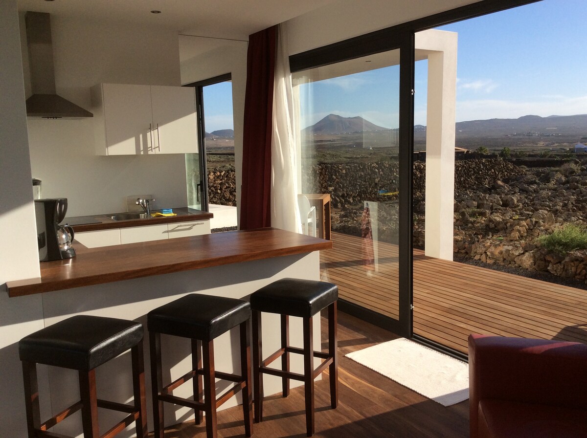 An open-concept kitchen area features a wooden countertop that faces large glass doors, allowing natural light to fill the space. Bar stools are positioned at the counter, and a view of the surrounding landscape and distant volcanic hills is visible through the doors.