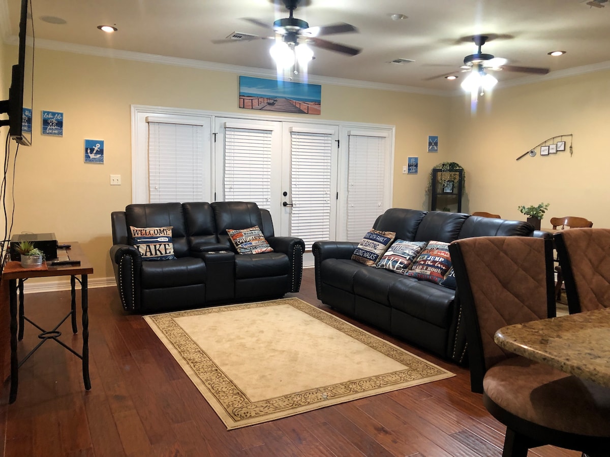 The living area features two comfortable black sofas adorned with decorative pillows. A warm area rug is placed centrally on the wooden floor, while natural light filters through the closed blinds of the glass door leading to the patio.