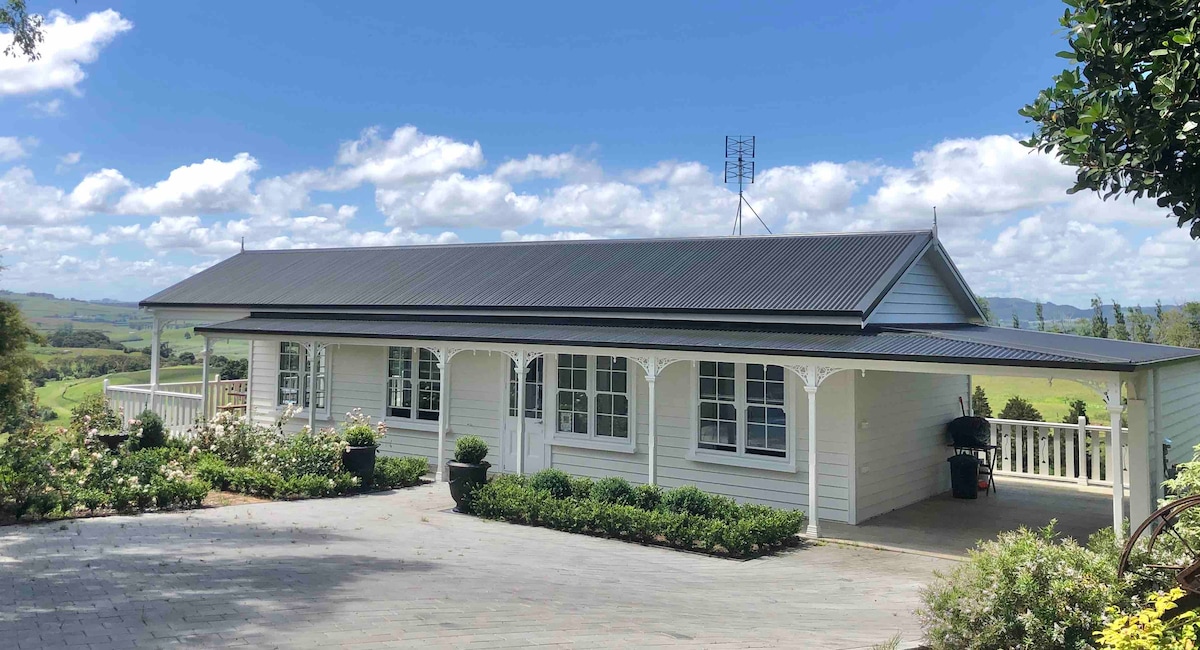 A newly constructed cottage is presented, featuring a large covered porch with decorative columns and numerous windows that allow for natural light. Lush greenery surrounds the exterior, providing a peaceful country atmosphere. Scenic views of the landscape are visible in the background.