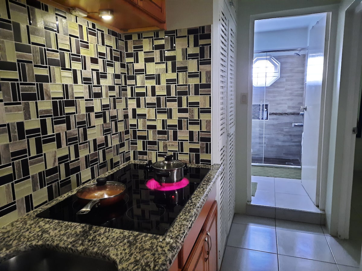 The kitchen features a modern stove set against intricately designed backsplash tiles in earthy tones. A pot simmers on the cooktop, complemented by wooden cabinetry. In the background, the entrance to a stylish bathroom can be seen, with neutral-colored tiles visible through an open door.