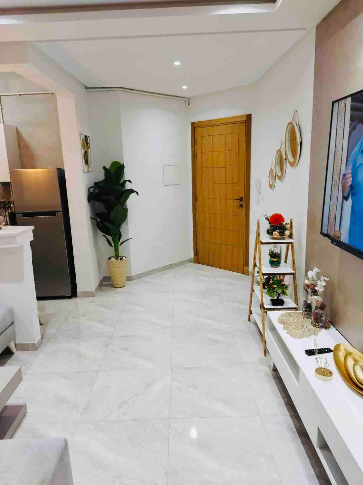 A modern hallway is visible, featuring smooth marble flooring and soft lighting from the ceiling. A sleek kitchen area is positioned to the left, while a decorative plant adds a touch of greenery. An inviting wooden door and wall art complete this stylish and airy space.