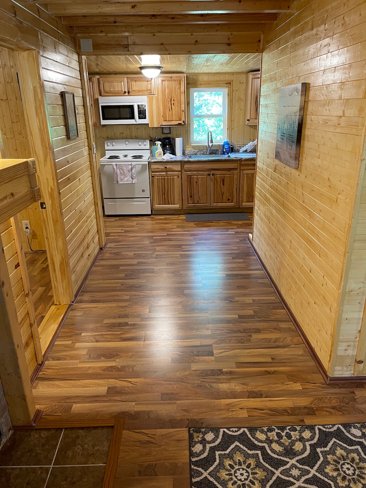 A well-lit hallway features warm wooden paneling and a polished floor, leading to a fully equipped kitchen on the right. The kitchen includes wooden cabinetry, a microwave, and a stove, with natural light filtering in through a window.