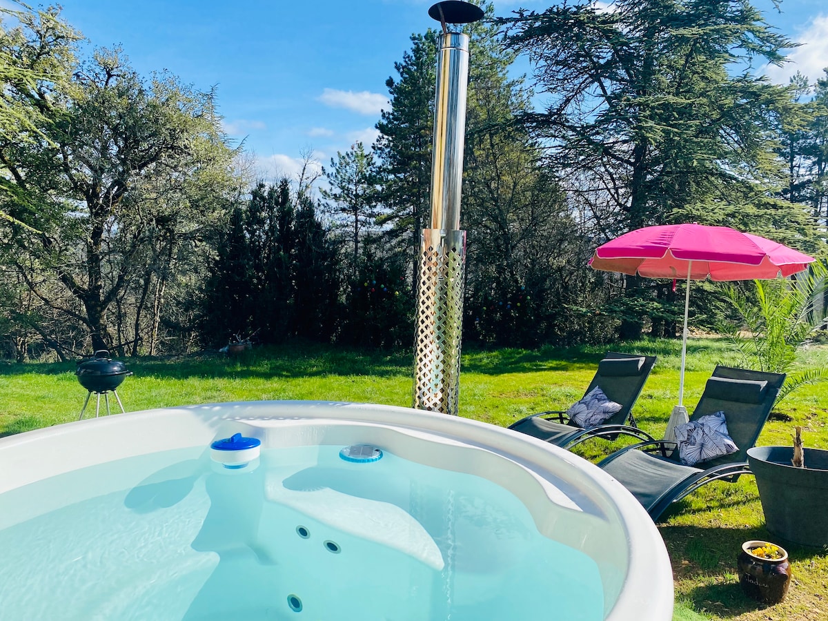 A private outdoor Nordic bath is framed by lush greenery, featuring a wood-burning stove. Two loungers with cushions complement a pink umbrella, and a small barbecue grill is nearby, inviting relaxation in nature.