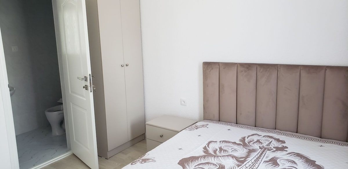 A bedroom is shown with a spacious bed featuring a light-colored headboard. A simple nightstand is positioned beside the bed. A white wardrobe is visible, and a private bathroom is accessible through a door in the background, showcasing tiled flooring.