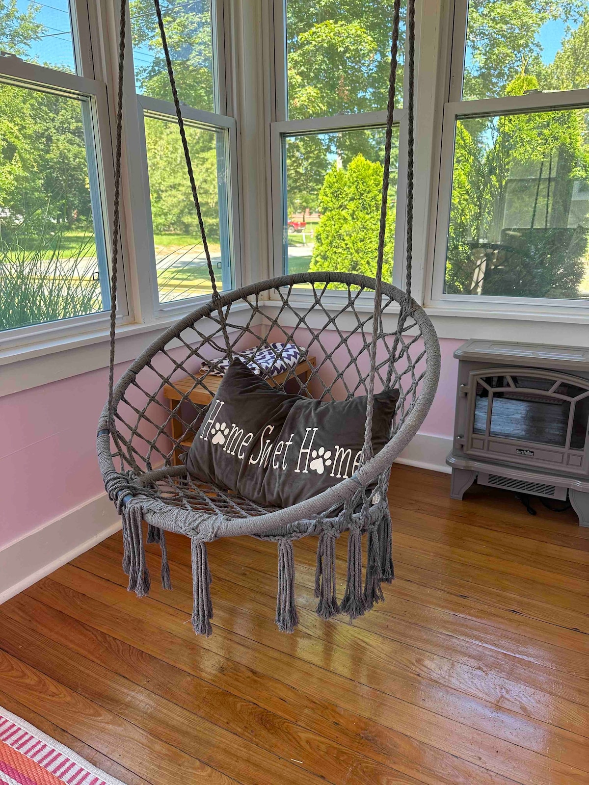 A cozy swinging chair is suspended in a sunlit corner. Two decorative pillows with the phrase 'Home Sweet Home' are placed within the chair. Surrounding windows allow for ample natural light, highlighting the warm wooden flooring and a small decorative heater nearby.