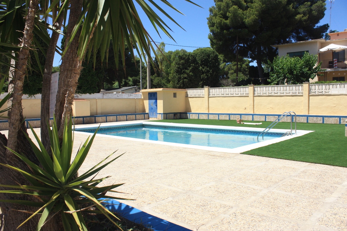 A community swimming pool is set within a landscaped area, featuring a clear blue water surface. Surrounding palm trees and green grass create a relaxing environment, while the pool deck provides ample space for sunbathing. A ladder leads into the water.