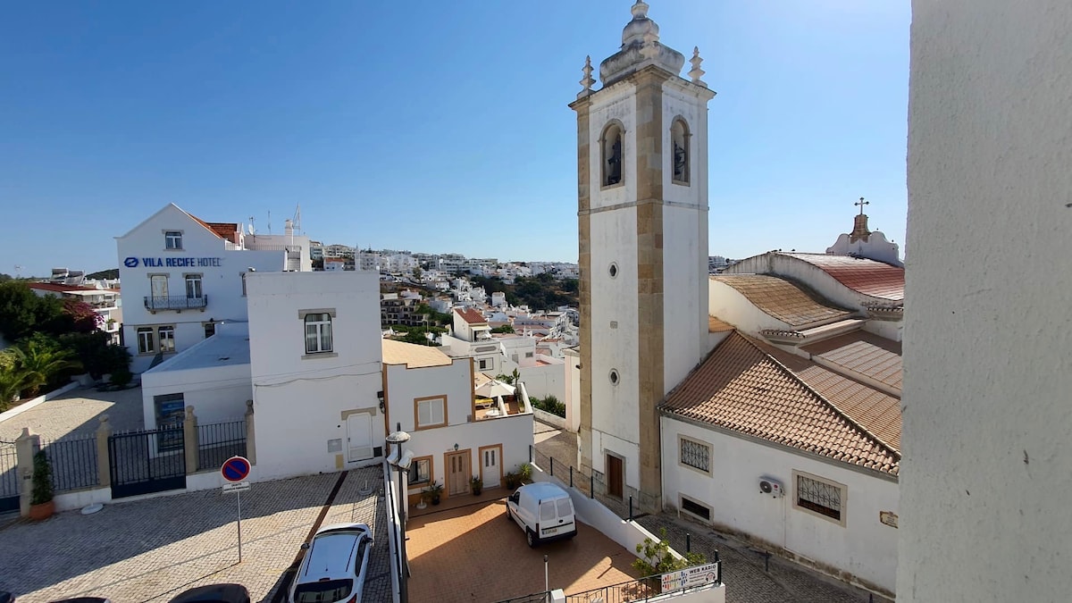 A view of the Old Town features a clock tower and a church, showcasing traditional architecture. Surrounding white buildings and a parked car are visible, with a clear blue sky enhancing the landscape. The area appears to be well-kept and pedestrian-friendly.