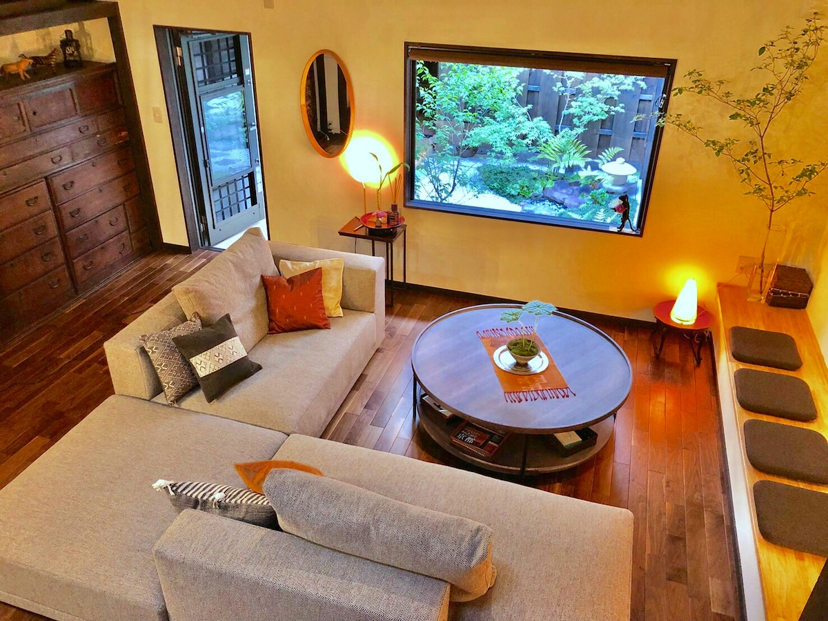 A spacious lounge area features a large, comfortable sectional sofa with accent pillows. A round coffee table sits in the center, elegantly presenting a decorative plant. A window offers a view of a serene Japanese garden, enhancing the atmosphere with natural light.