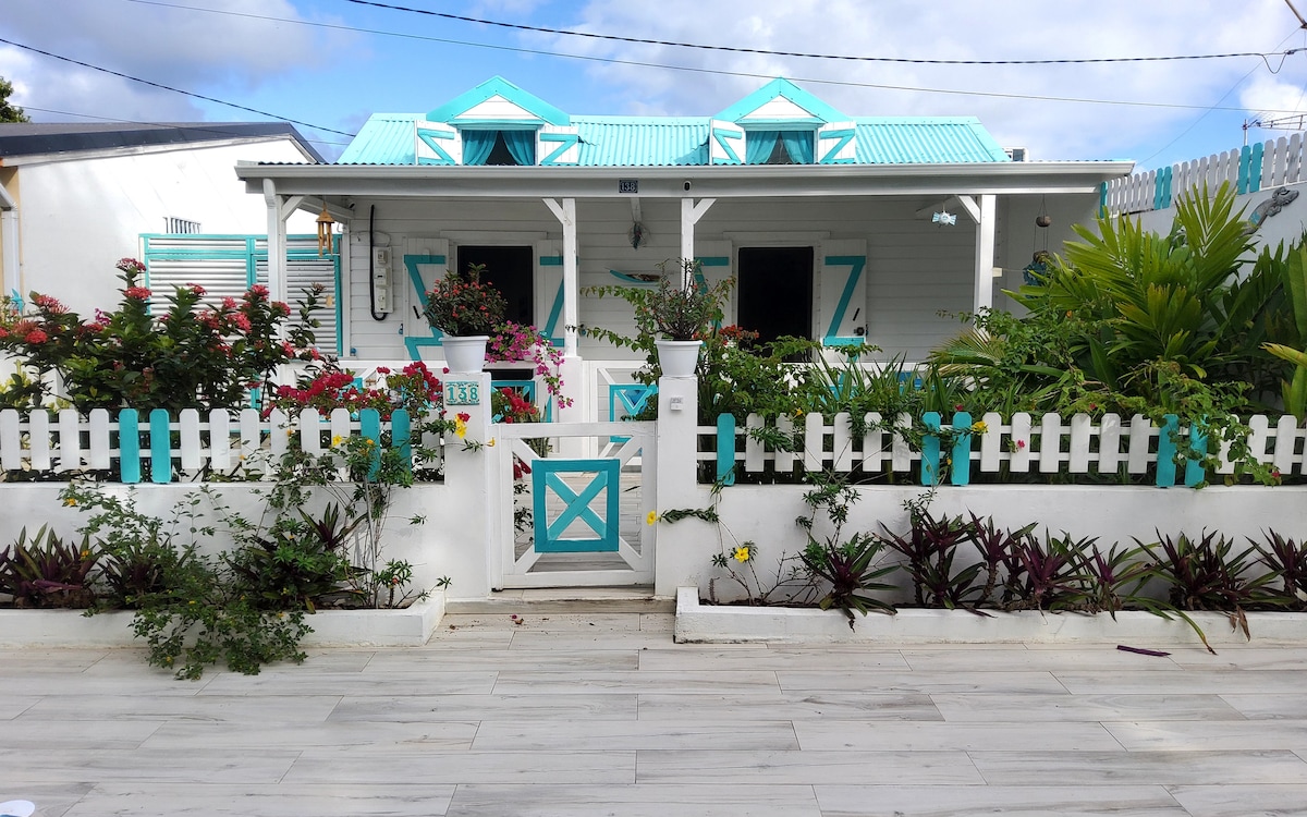 A charming creole house is framed by vibrant foliage and a white picket fence, displaying colorful shutters and a light blue roof. The welcoming entrance is adorned with a garden featuring blooming flowers and lush greenery, presenting a picturesque setting in the tropical landscape.