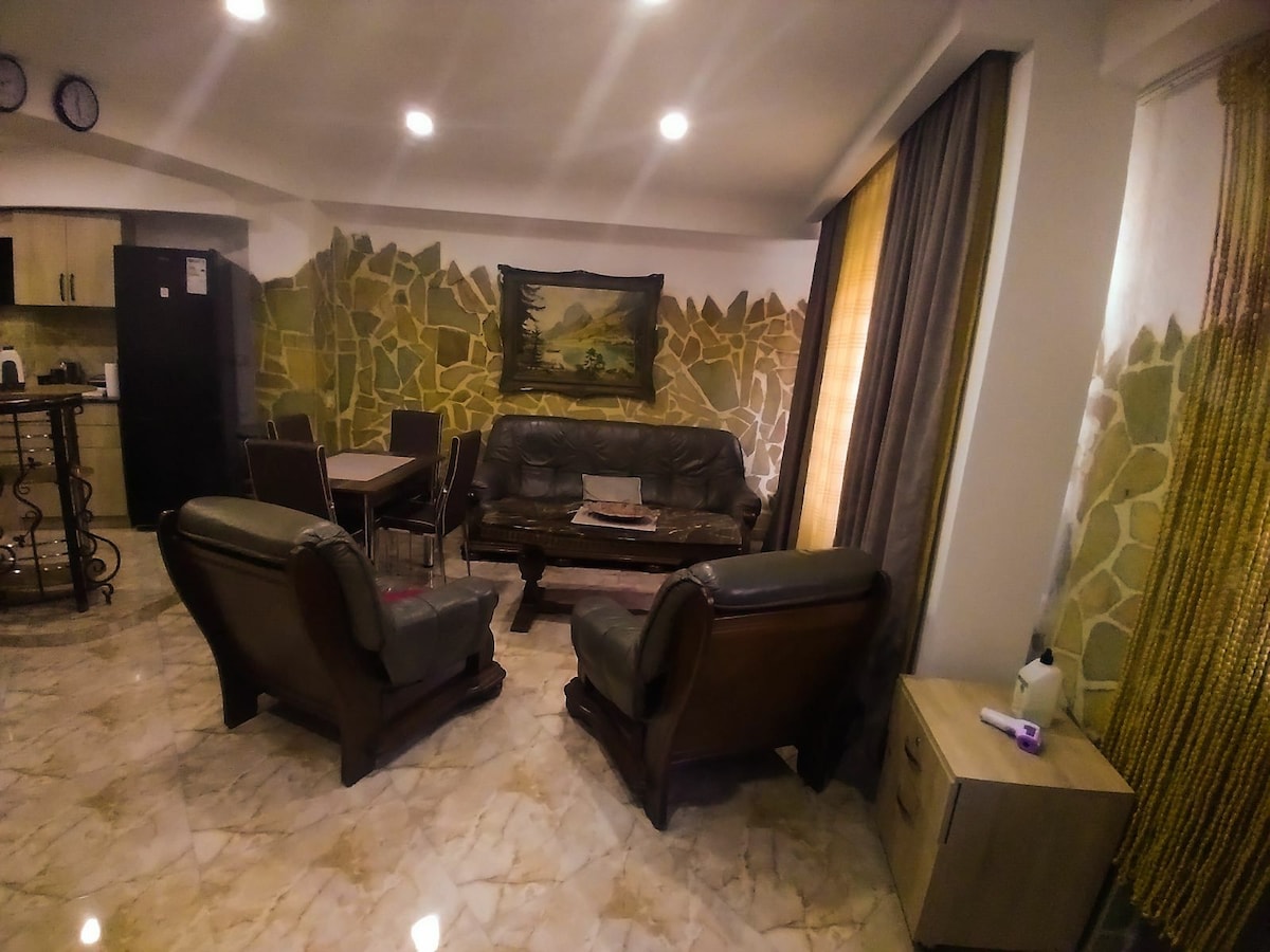 A living area is defined by a leather sofa and two armchairs, surrounded by stone-patterned walls. A wooden dining table is visible nearby, with a modern kitchen area in the background. Natural light filters in through floor-to-ceiling curtains.