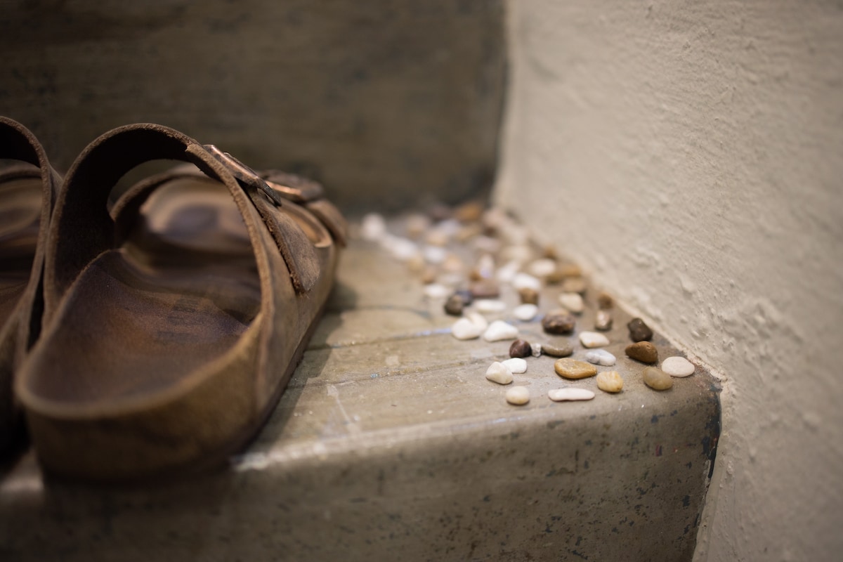 A pair of brown sandals rests on a light-colored staircase, next to a collection of smooth, multicolored pebbles scattered across the step, adding a natural element to the space.