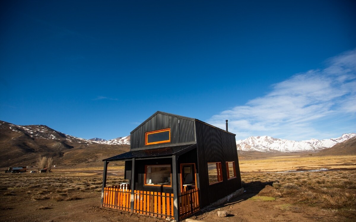 A modern black cabin with orange accents is set against a backdrop of the Andes mountains. The spacious porch features a railing, and large windows allow for natural light. The surrounding landscape includes grassy fields and distant snow-capped peaks under a clear blue sky.