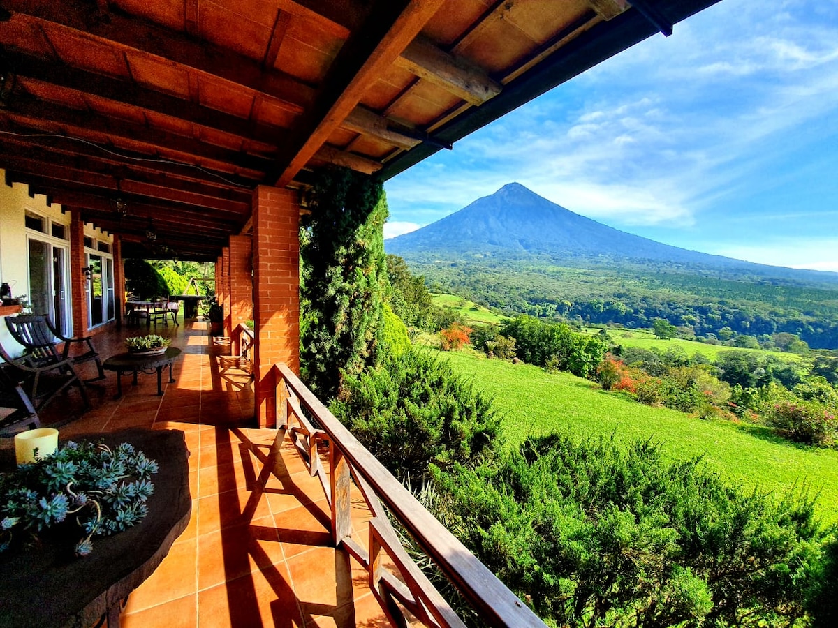 A spacious porch features wooden beams and a tile floor, offering a view of a prominent volcano. The surrounding landscape is filled with lush greenery, while comfortable chairs provide a place to relax and take in the scenery.