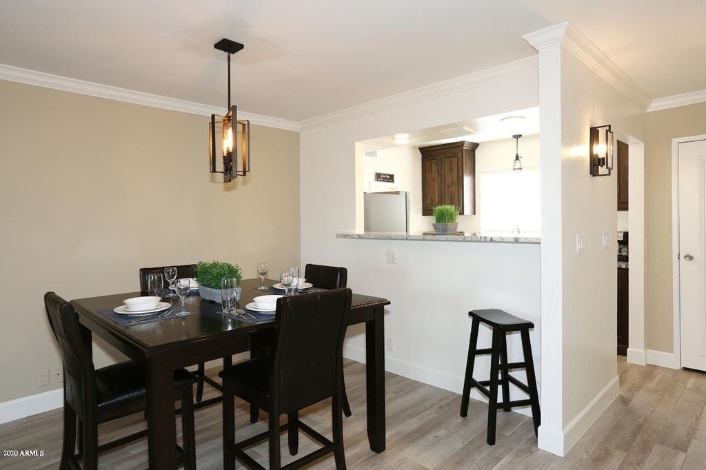 A dining area is highlighted by a dark wood table set for four, surrounded by matching chairs. A black stool is positioned at the edge, and a small plant adds a touch of greenery on the table. An open kitchen space is visible in the background.