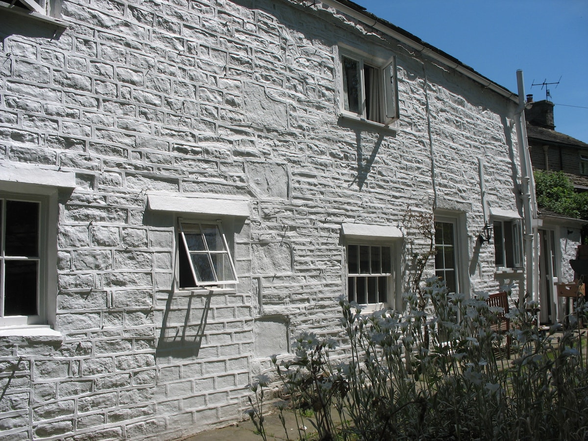 The exterior of a stone-built cottage is visible, featuring a textured whitewashed facade. Several windows, including one small paneled window, create an inviting appearance. A modest garden area with greenery and outdoor seating is located adjacent to the cottage, enhancing the overall charm of the property.