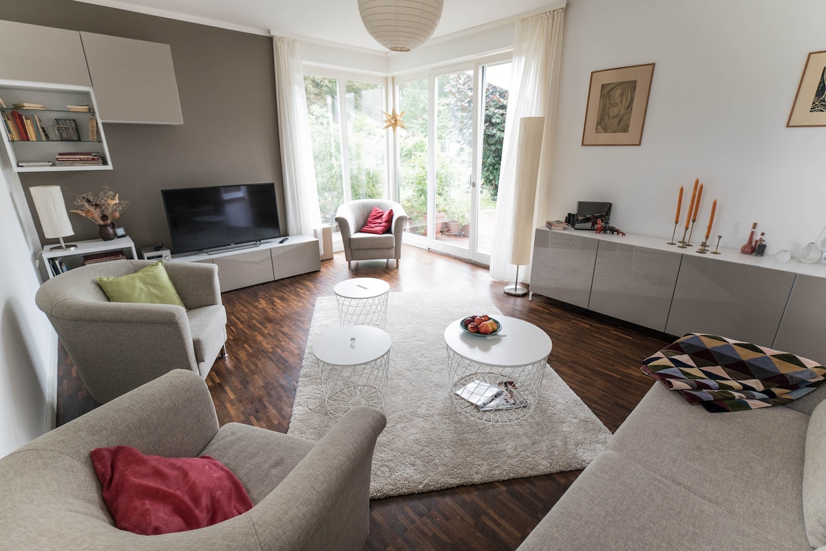 A light-filled living area contains two soft sofas and two accent chairs arranged around a small coffee table. A television is positioned on a low cabinet against one wall. Large windows offer views of the garden, with soft curtains allowing natural light to fill the space.