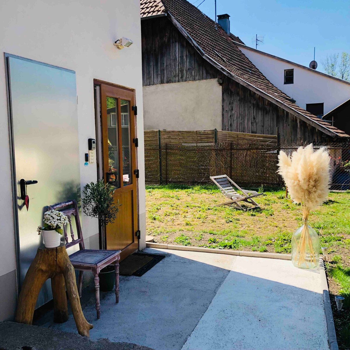 The entrance features a wooden door beside a glass panel. A rustic chair and a decorative plant are positioned on a small patio area. Pampas grass in a clear vase adds a natural touch, while a sun lounger is visible in the grassy outdoor space.