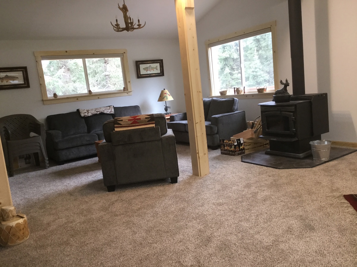 A spacious living area is presented with two gray sofas and a single armchair arranged around a central coffee table. A wood-burning stove stands in the corner, and large windows allow light to filter in, providing views of the surrounding trees.