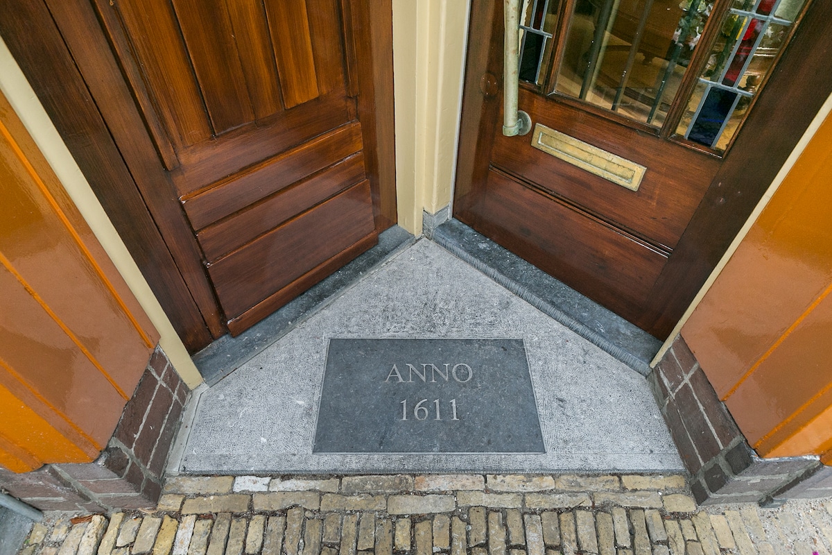 The entrance features two wooden doors, one with decorative glass panels. A stone plaque inscribed with 'ANNO 1611' is set in the ground between the doors, adding historical significance. The surrounding area is made from cobblestones, enhancing the welcoming atmosphere.