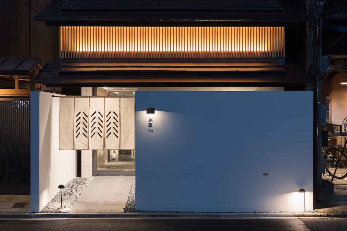 The entrance showcases a modern architectural design, featuring a white exterior wall and warm wooden accents. Soft lighting illuminates the entrance, where decorative curtains hang. A pathway lined with small stones leads towards the entrance, enhancing the minimalist aesthetic.