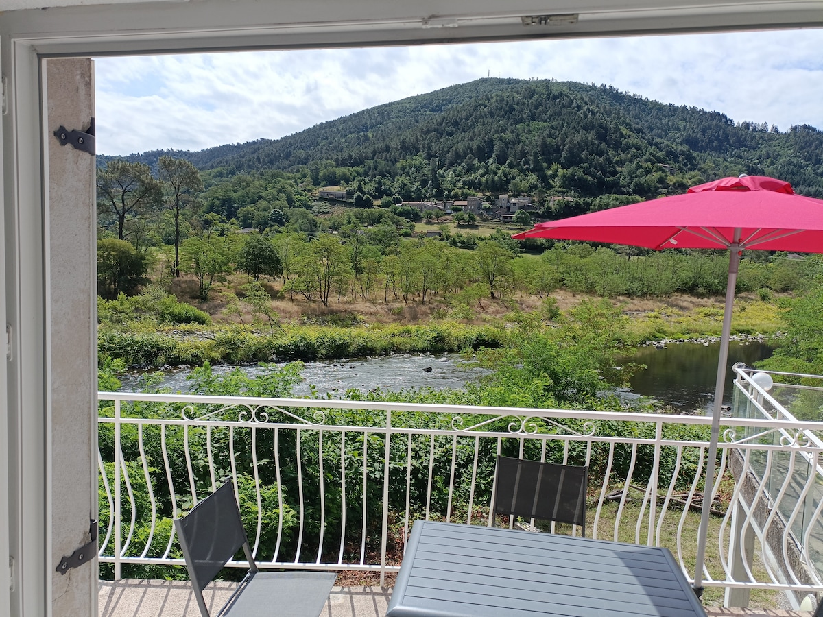 A view from an open door reveals a scenic landscape featuring a lush hillside and a flowing river. A small table and chairs sit on the terrace, complemented by a bright red umbrella that provides shade.