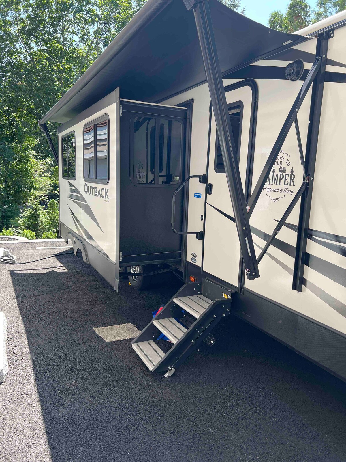 An outdoor view of the RV shows its sleek exterior with a prominent Outback emblem. A canopy extends from the side, providing shade over the entrance. Sturdy steps lead up to the door, which is flanked by large windows that allow natural light to enter.