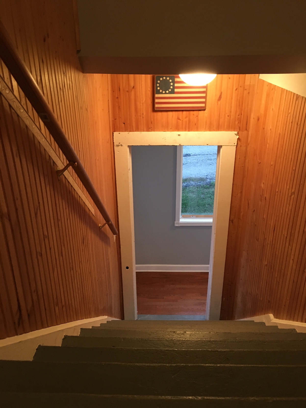 An open staircase leading downwards is shown, with wooden paneling on the walls. A small window illuminates the space, and a flag is displayed above the doorway at the bottom of the stairs. The steps are painted a muted color, enhancing the homey feel.
