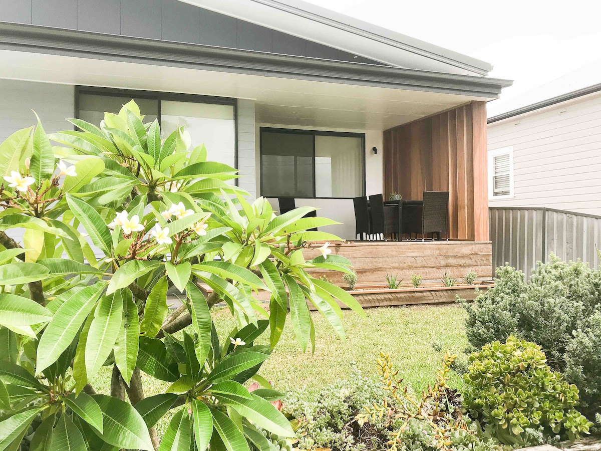 The entrance is framed by lush green foliage and flowers, leading to a large front deck. Outdoor seating is visible, creating a welcoming space for relaxation. The two-story home features large windows that allow natural light into the interior.