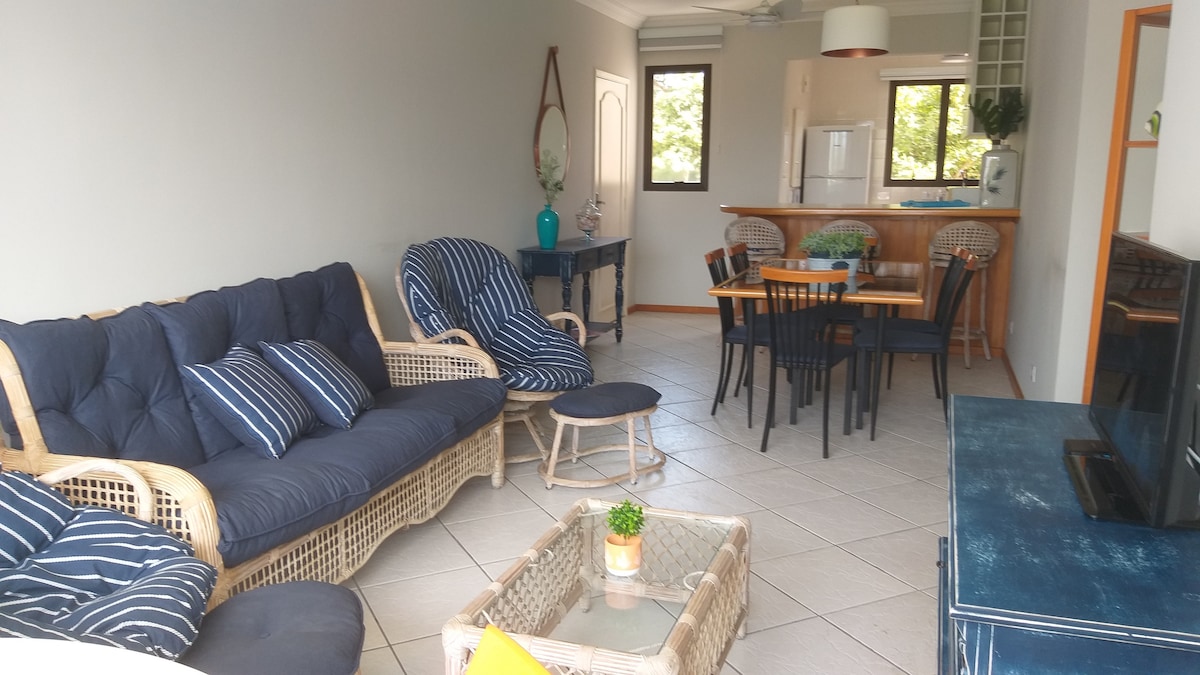A living area is featured with a cozy seating arrangement including a rattan couch and armchairs, complemented by striped cushions. A dining table for six is visible beside a kitchenette with a bar. Large windows allow natural light to brighten the space.