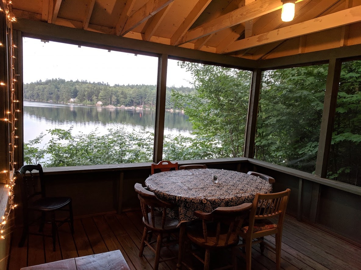 A screened porch features a round dining table surrounded by four wooden chairs, offering a serene view of the calm lake and lush greenery. Soft lighting hangs above, illuminating the space while the natural surroundings create a tranquil atmosphere.
